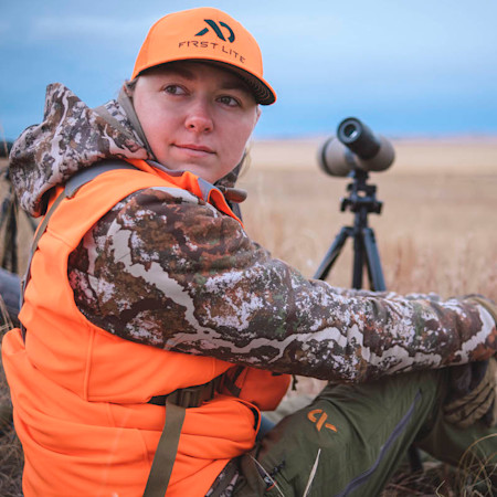 Woman wearing FIRST LITE cap and orange vest seated with spotting scope in field
