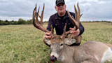 Man kneeling behind large buck, holding its antlers in a grassy field