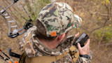 Hunter in camo cap and jacket checks phone in treestand; 'FIRSTLITE' on hood