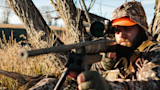 Hunter aiming a scoped rifle from low position by a tree, wearing camouflage and orange cap