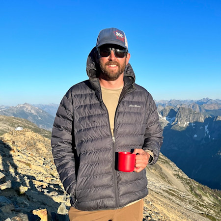 Man on mountain ridge holding red mug, wearing sunglasses and a cap reading "30L"