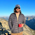 Man on mountain ridge holding red mug, wearing sunglasses and a cap reading "30L"