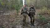 Two camouflaged hunters walking through leaf-strewn woods, one carrying a shotgun