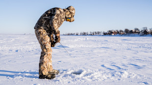 Man ice fishing on frozen lake, wearing camouflage jacket and mirrored goggles