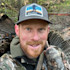 Man with red beard carrying a turkey, wearing camouflage and a cap labeled "HuntStand"