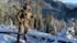 Hunter in camouflage holding a compound bow on a snowy mountain ridge overlooking a forested valley