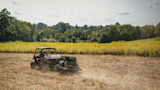 UTV towing a seed drill across a harvested field, kicking up dust toward a tree-lined meadow