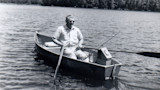 Older man in sunglasses rowing a small boat on a lake, fishing rod and tackle box onboard