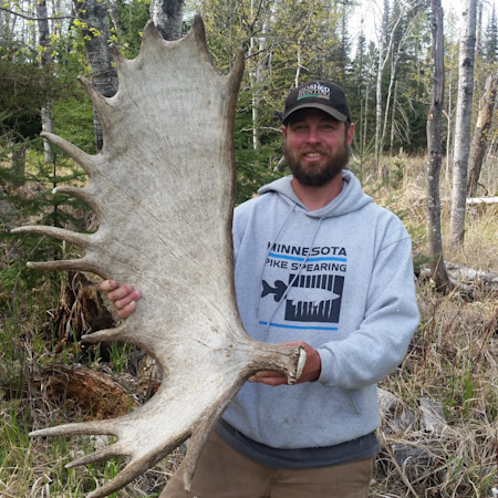 Man holding large moose antler in woods, wearing hoodie reading "MINNESOTA PIKE SPEARING"