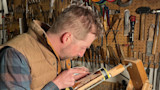 Man carving checkered gunstock clamped in a vise in a workshop with pegboard tools