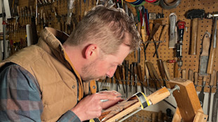 Man carving checkered gunstock clamped in a vise in a workshop with pegboard tools