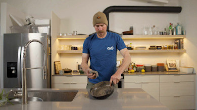 Man cleaning cast-iron skillet at kitchen counter, wearing blue shirt reading Cal's