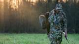 Hunter in camouflage carries turkey over shoulder, shotgun in hand, binoculars around neck in sunset field