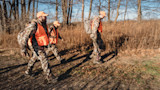 Three hunters walking through autumn grass wearing orange safety vests and camouflage gear