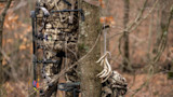 Hunter in camouflage holding a compound bow; shed antlers hanging from a tree