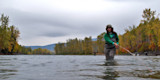 Woman wading chest-deep casting a fly rod in a river flanked by autumn trees