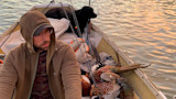 Man in hooded jacket and glasses sitting in small boat with black dog, shotgun, and three pheasants