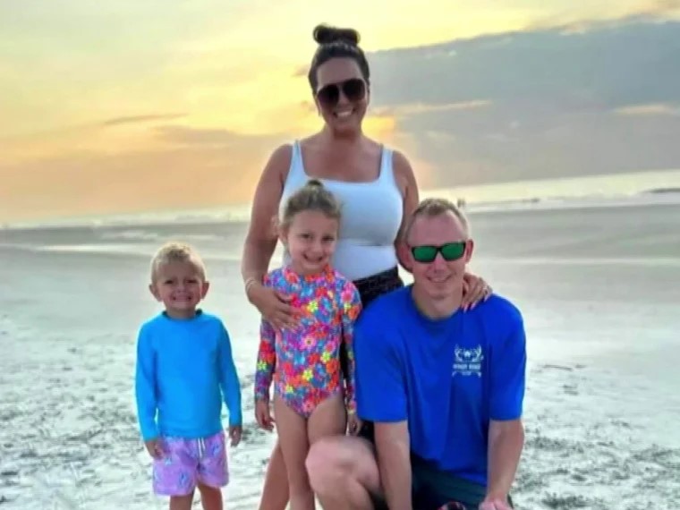 Family of four smiling on sandy beach at sunset: woman standing behind girl, boy, and kneeling man.