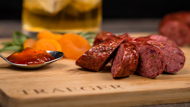 Sliced dark red sausage with red jam on spoon and apricot slices on wooden board 'TRAVELER'