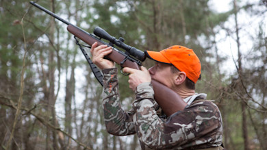 Hunter aiming scoped rifle upward in woods, wearing orange cap and camouflage