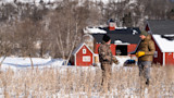 Two hunters talking in a snowy field with a red barn in the background