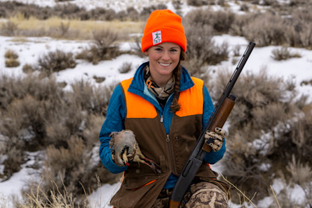 Smiling woman hunter in orange beanie with 'TRCP' patch, holding shotgun and chukar in snowy sagebrush