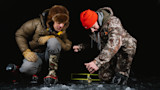 Two hunters kneel on black ice tending a yellow tip-up, one in an orange beanie