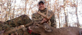 Hunter holding large-antlered buck with blood on its mouth, smiling in leaf-strewn woods