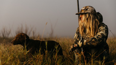 Hunter crouching with shotgun next to brown retriever in marsh grass