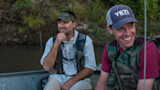 Two fishermen in a boat on a river; right man smiling wearing cap reading YETI COOLERS