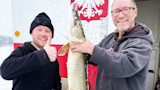 Two men holding a large muskie at an ice shack painted with a red eagle and '2021'