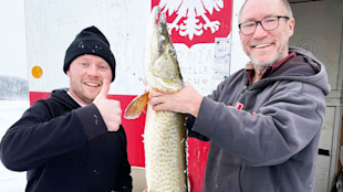 Two men holding a large muskie at an ice shack painted with a red eagle and '2021'