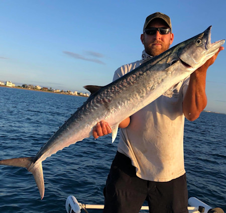 Fisherman holding large mackerel on boat; shoreline houses in background