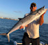 Fisherman holding large mackerel on boat; shoreline houses in background