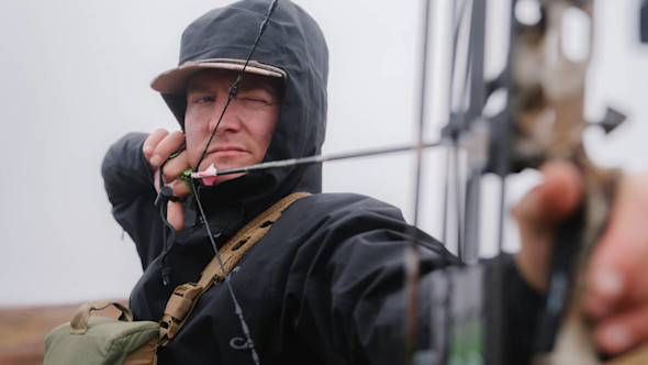 Hunter in hooded jacket drawing a compound bow, pink-nocked arrow at mouth