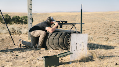 Man kneeling, aiming a scoped rifle from a tire rest on open prairie