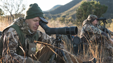 Two hunters seated in grass; foreground hunter looking through a spotting scope on tripod