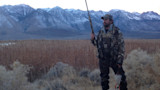 Hunter holding shotgun and two ducks in marsh with snow‑capped mountains in background