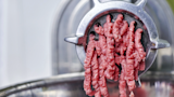 Raw ground meat being pushed through a metal meat grinder into a stainless bowl