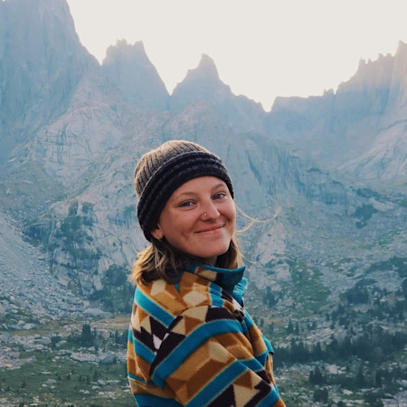 Smiling woman in knit beanie and geometric-pattern fleece with alpine peaks behind