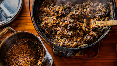 Cast-iron pot of rotini pasta with shredded meat and a small pot of beans on wooden table