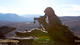 Hunter seated on ridge looking through a spotting scope on tripod, backpack and rifle nearby