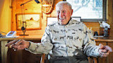 Elderly man laughing in wood-paneled cabin, wearing button-down shirt patterned with elk