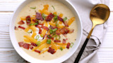 Creamy cauliflower soup topped with cheddar, bacon, green onions and sour cream, gold spoon nearby