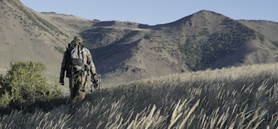 Hunter carrying backpack and compound bow walking through tall grass toward distant mountains