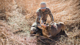 Hunter kneeling beside harvested buck with antlers in dry grass