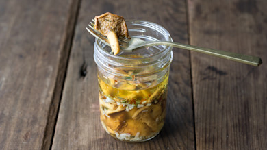 Jar of pickled wild mushrooms, fork holding one mushroom above jar on wooden table