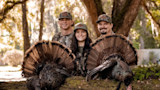 Three hunters in camouflage caps smiling behind two harvested wild turkeys with fanned tails on a log