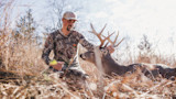 Man kneeling in dry grass, hand on a harvested buck's antlers