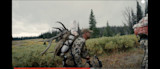 Hunter carrying elk antlers and wrapped meat on backpack, holding bow in alpine meadow
